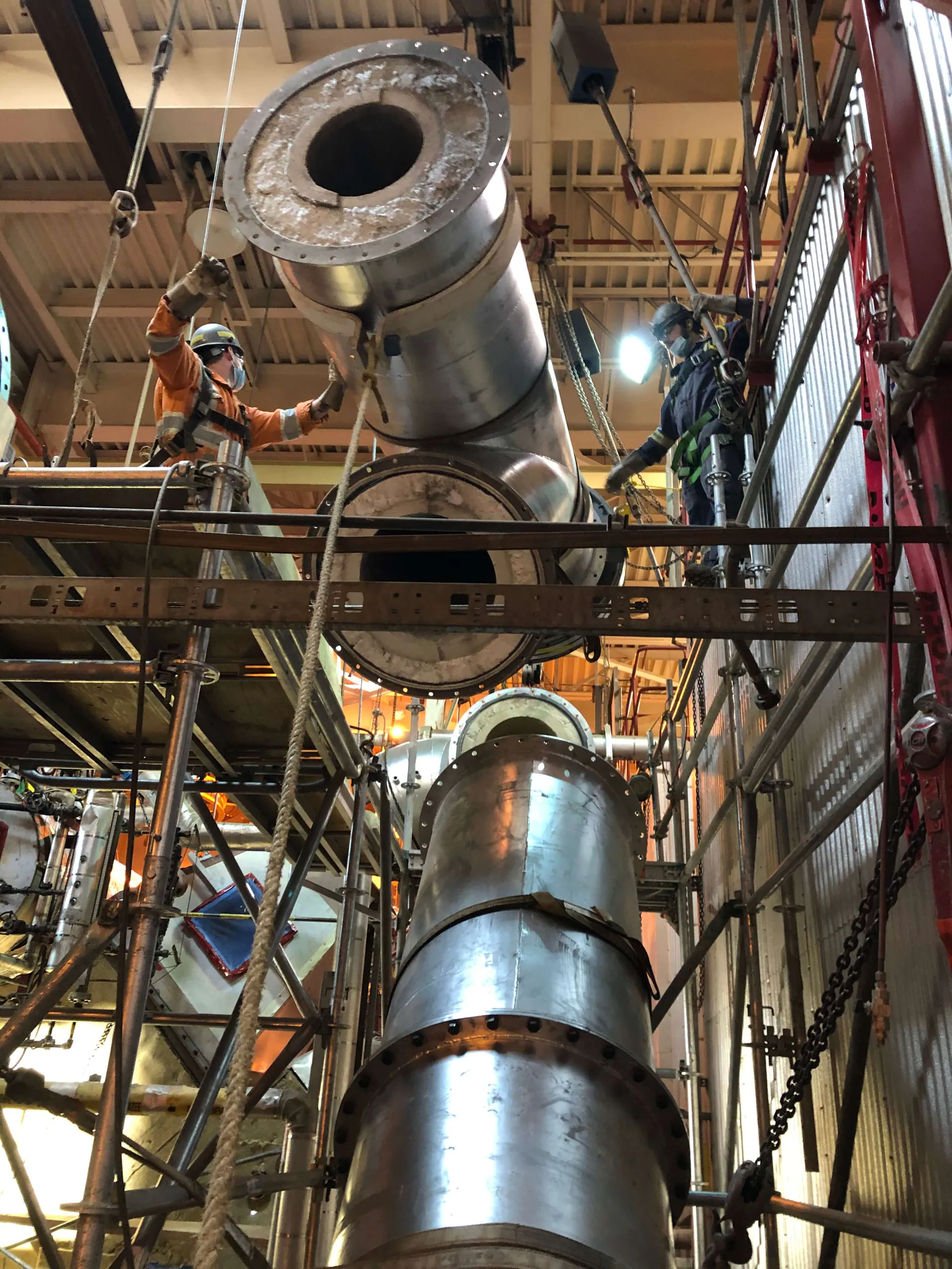 PPE-clad workers guide a large refractory-lined industrial duct elbow into position within scaffolding.