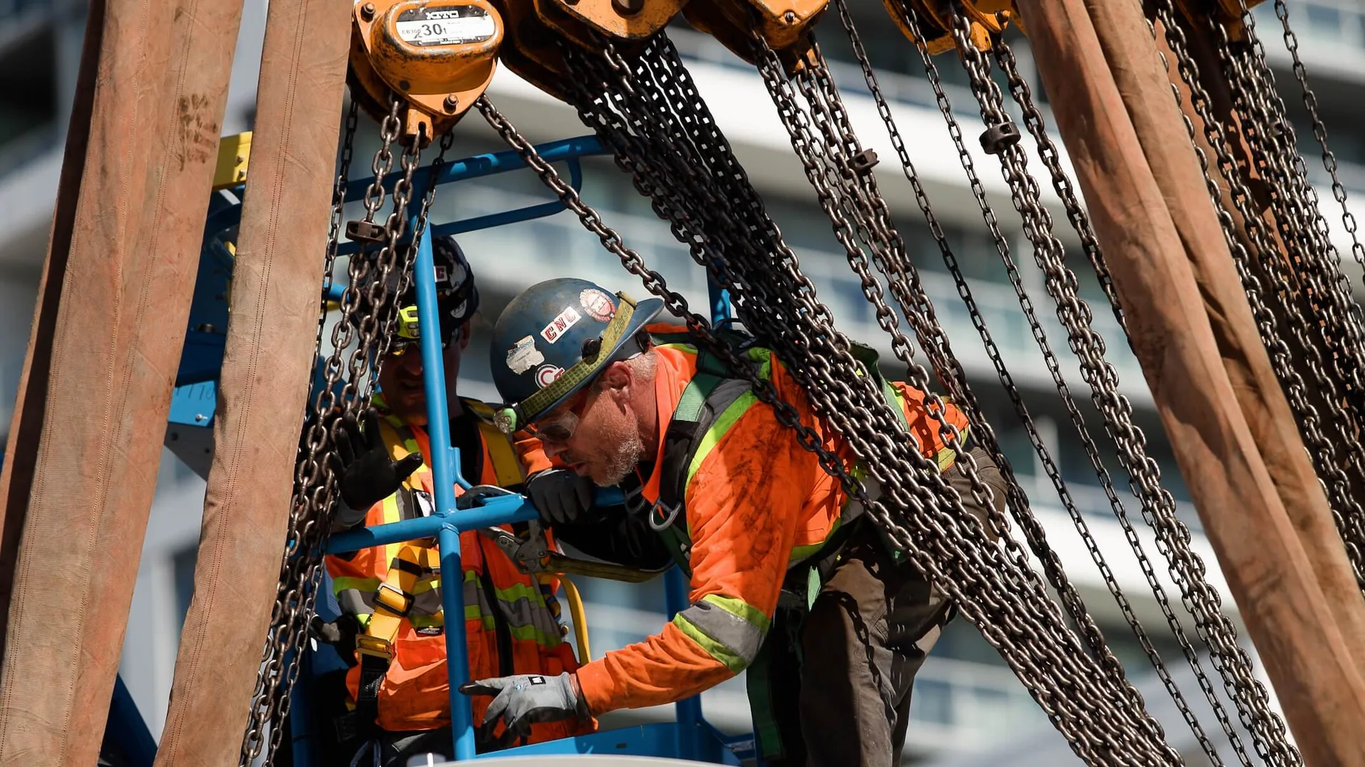 Close view of a hard-hatted worker in fall protection operating among chain hoists and lifting slings.