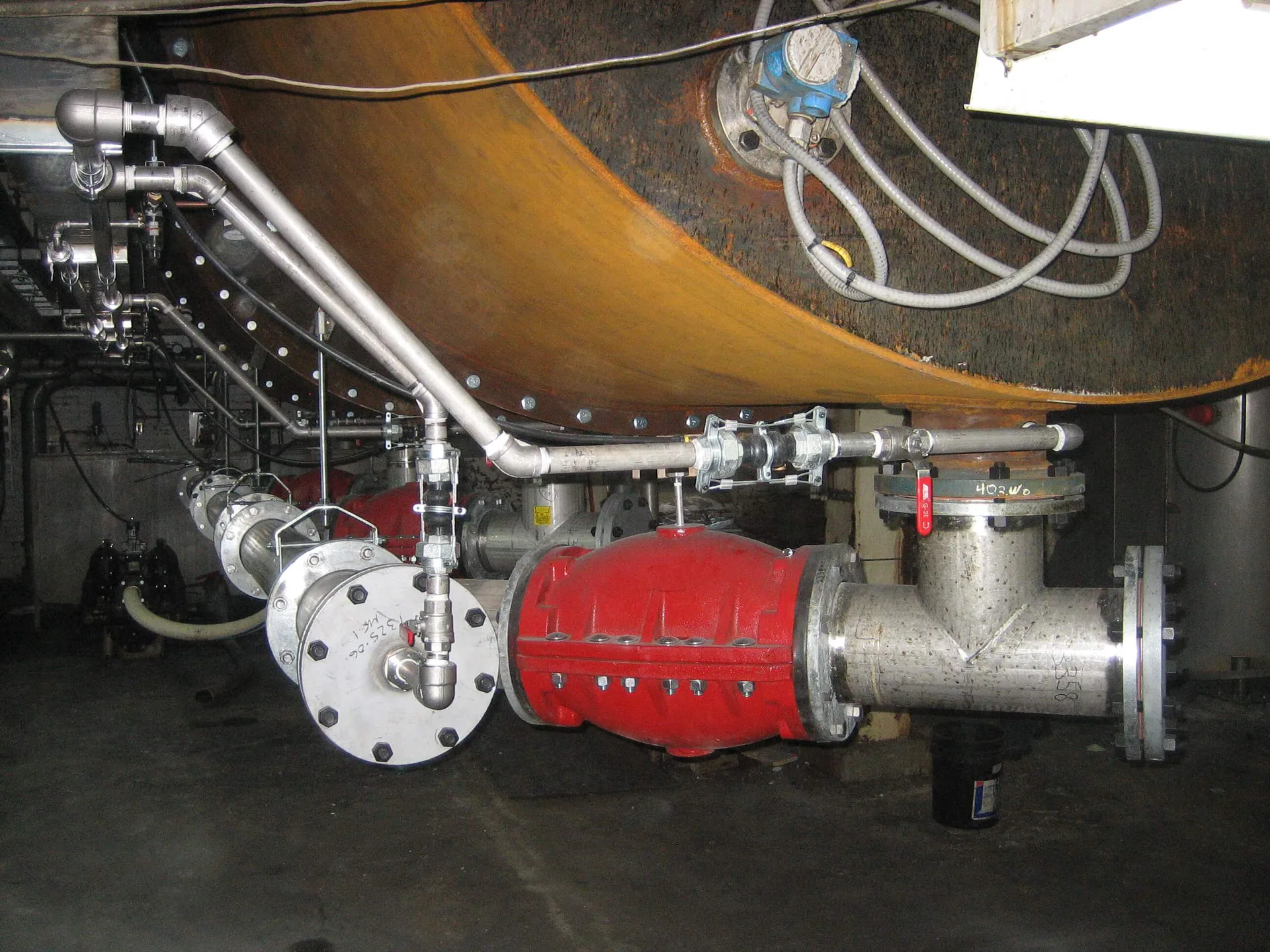 Stainless industrial pipe manifold with valves and a large flanged connection beneath a cylindrical surface.