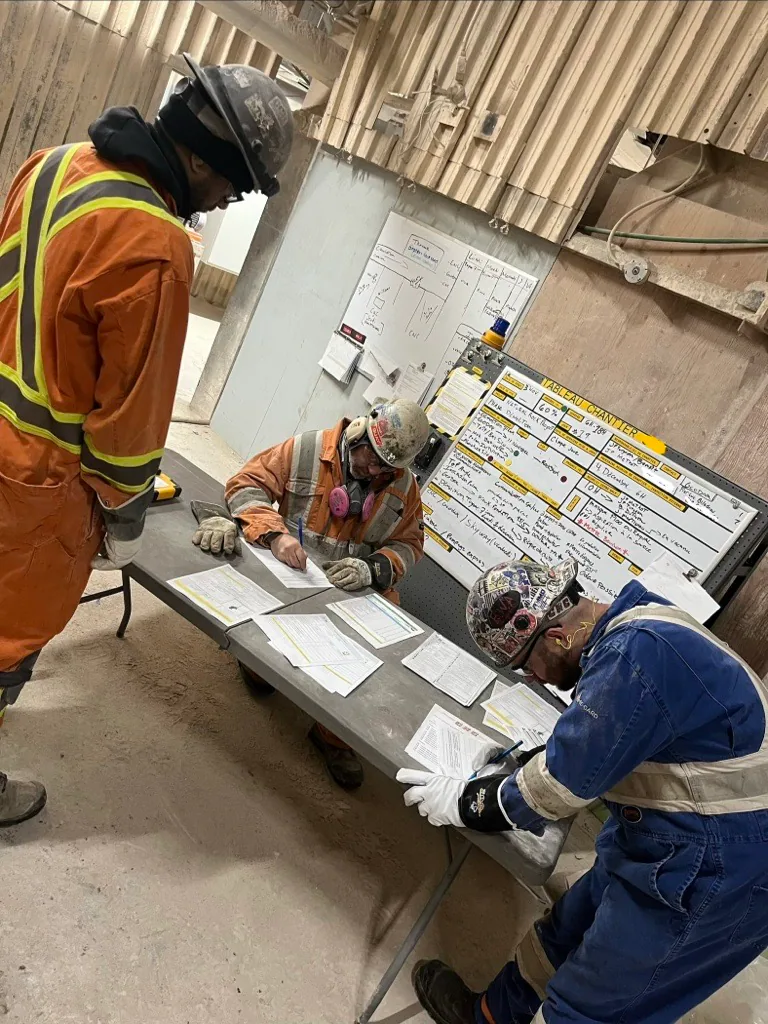 Three PPE-clad workers complete paper forms at a table beside a whiteboard or job board.