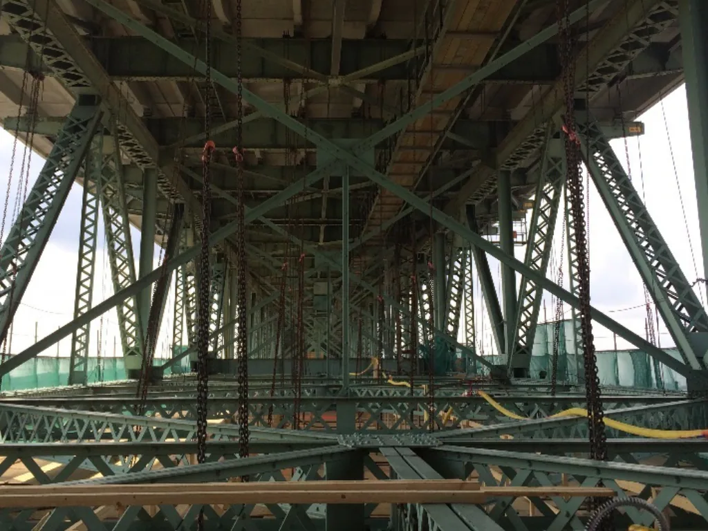 Underside of a bridge steel frame with chain hoists and protective netting.
