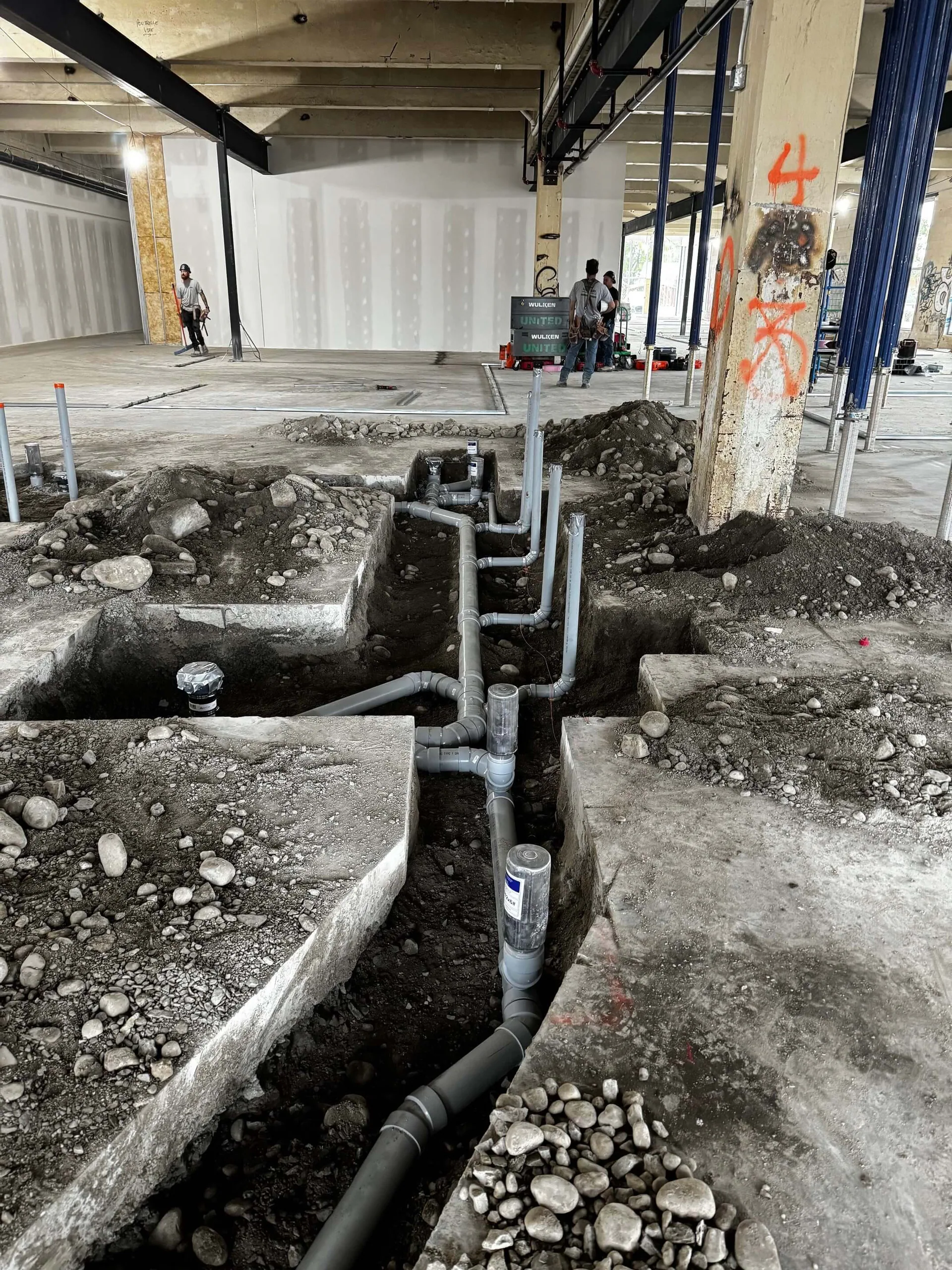 Exposed below-slab grey drain piping in trenches during a commercial interior renovation, with workers in the background.