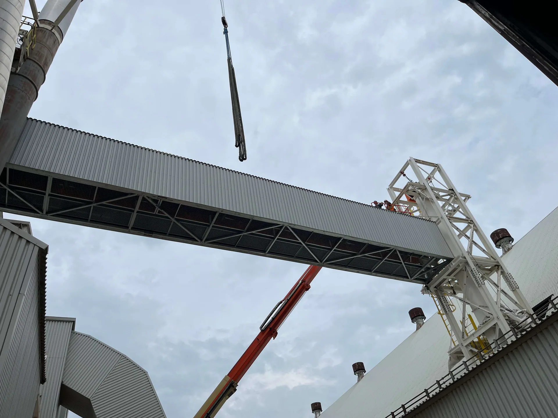Enclosed steel gallery installed between two industrial buildings, with a crane and two workers on top.