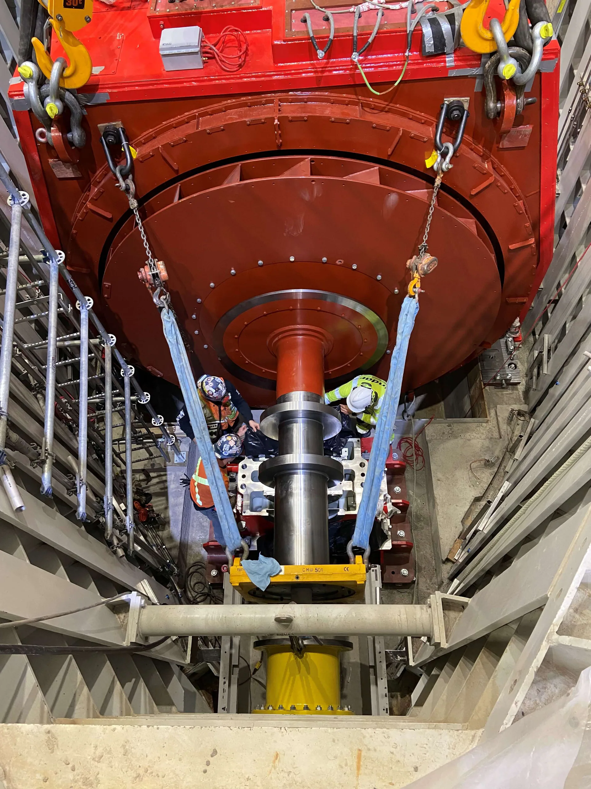 Top-down view of workers beside a massive red rotor or generator assembly and polished shaft in a powerhouse.