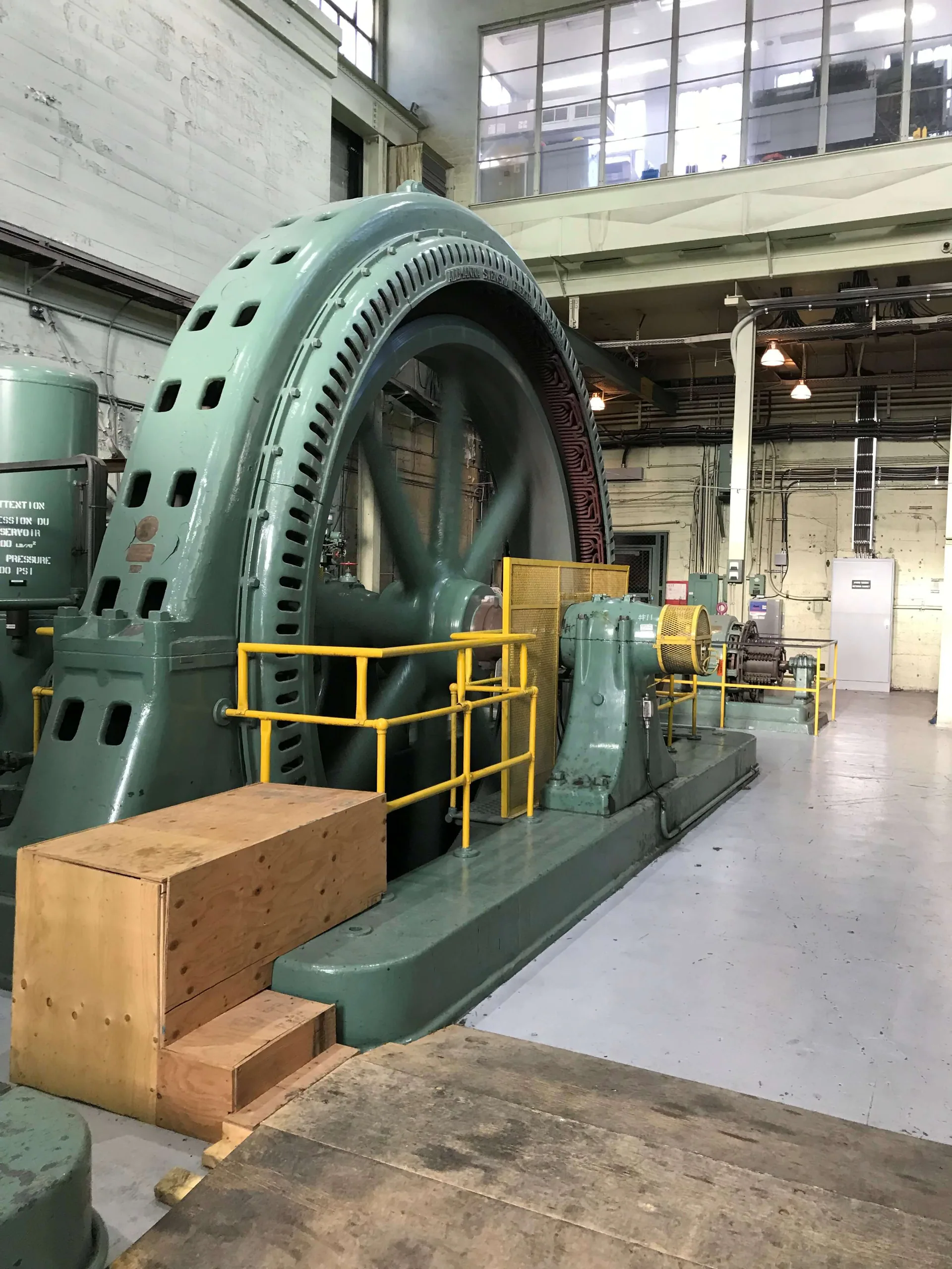 Large green historic generator or rotating machine displayed inside a powerhouse with yellow perimeter guards.