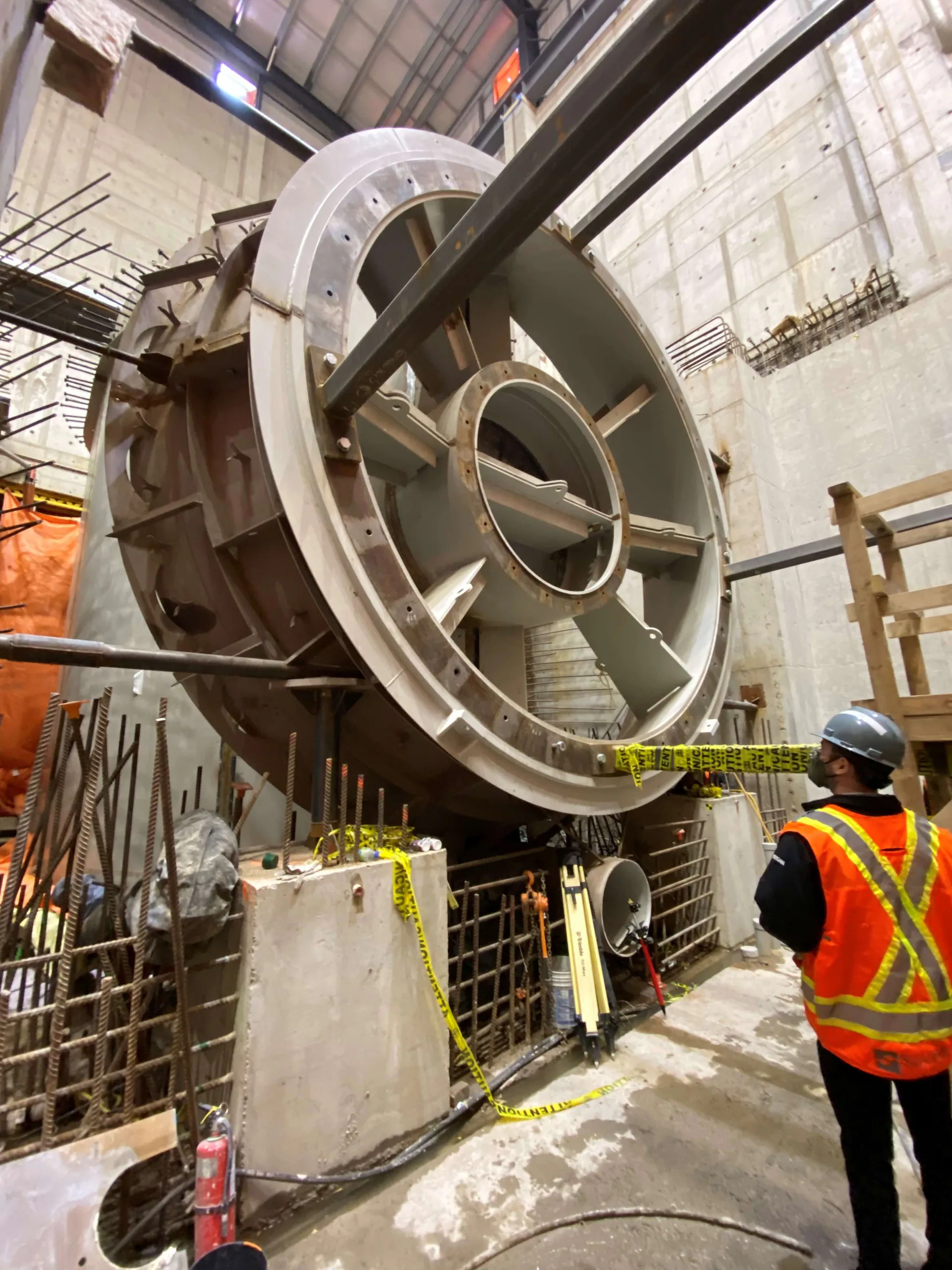 Large circular steel machine housing being positioned in a concrete powerhouse while a worker observes.