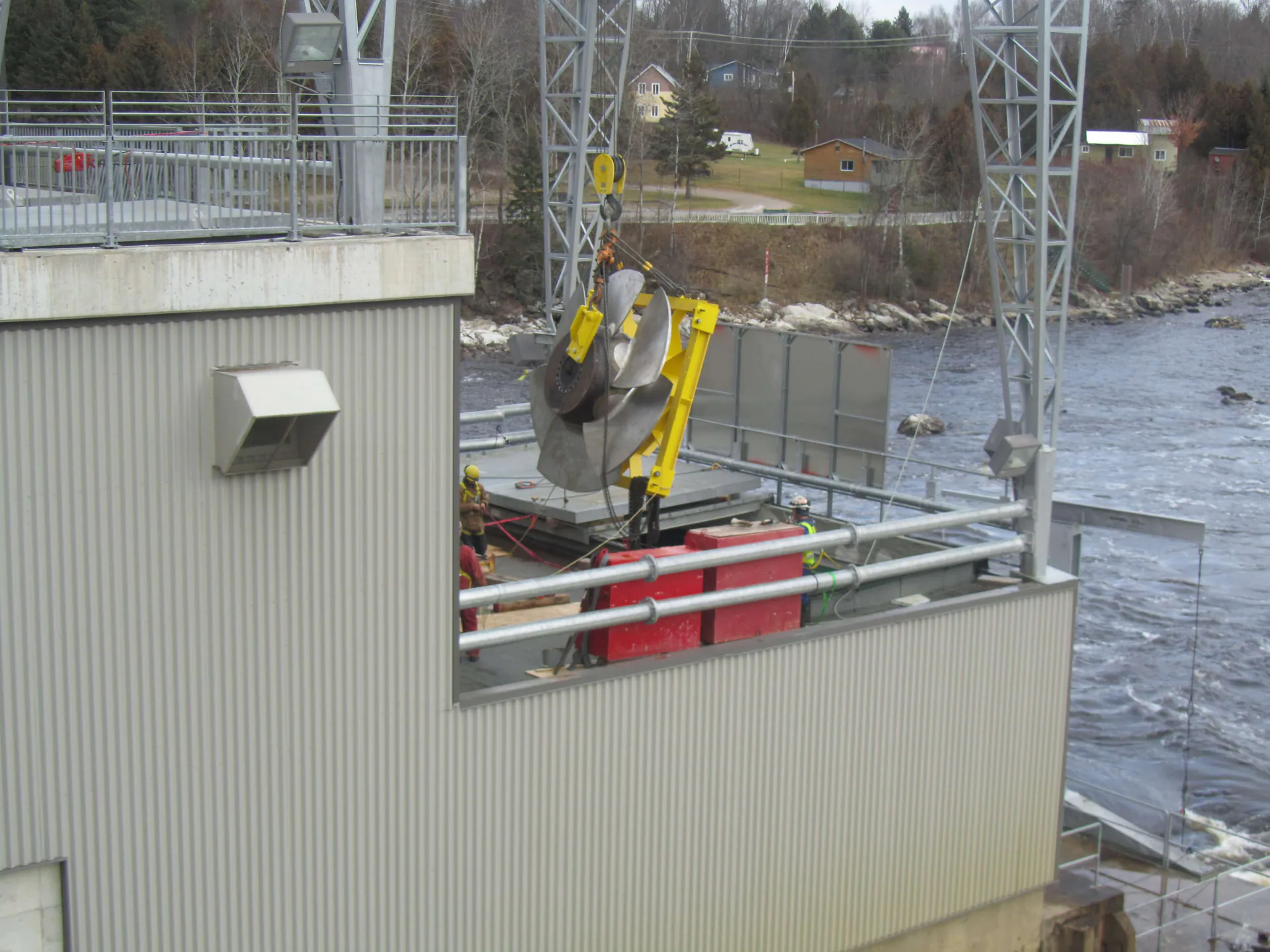 Large silver turbine-runner-like component is hoisted outdoors near a river while workers stand nearby.