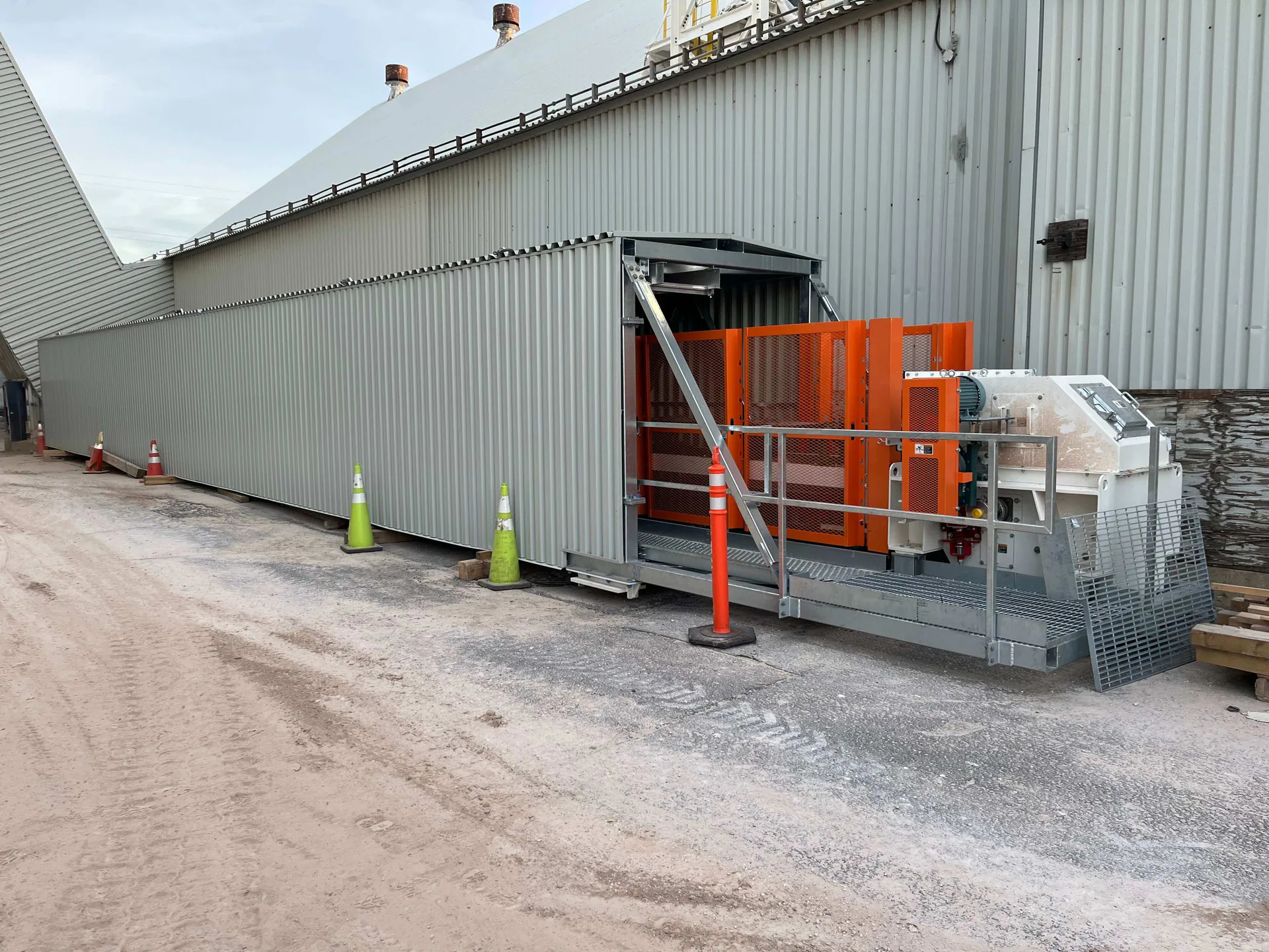 Completed enclosed steel gallery at ground level beside an industrial plant, with orange guarded drive or mechanical equipment at one end.