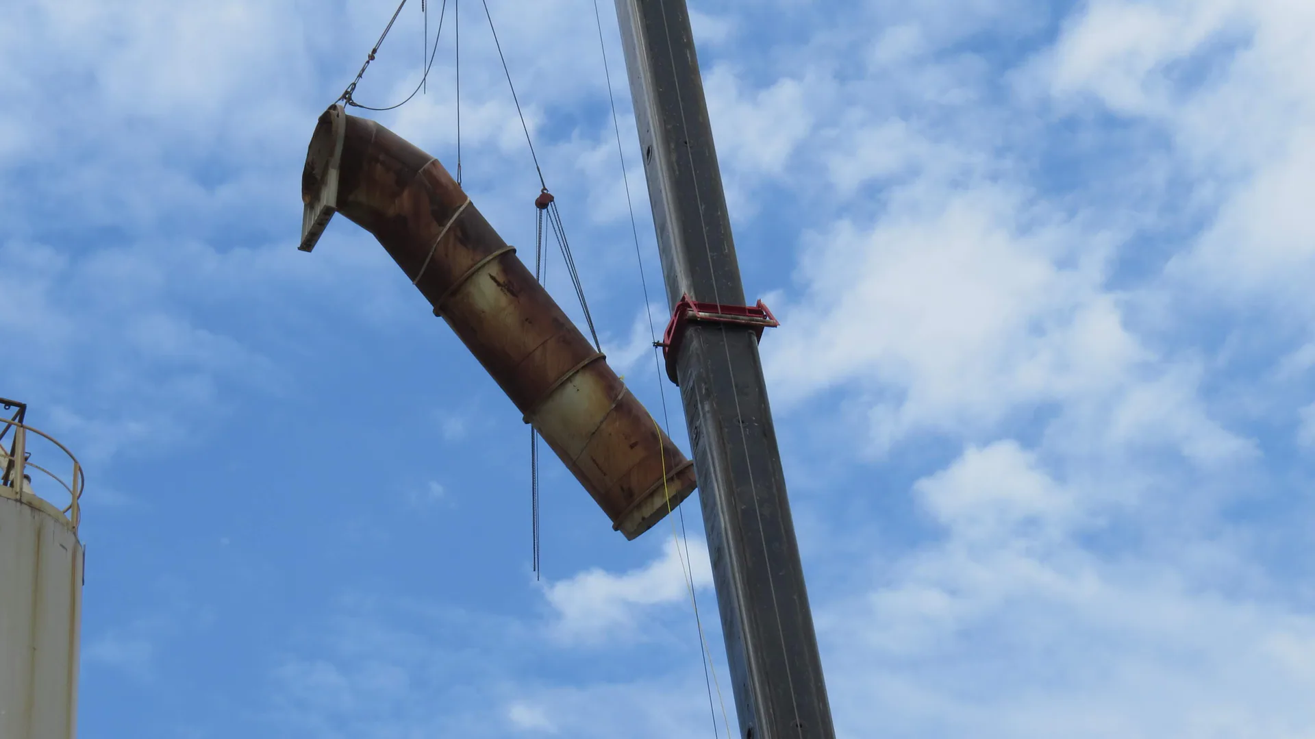 Large curved weathered industrial duct or shell section is suspended from a crane against the sky.