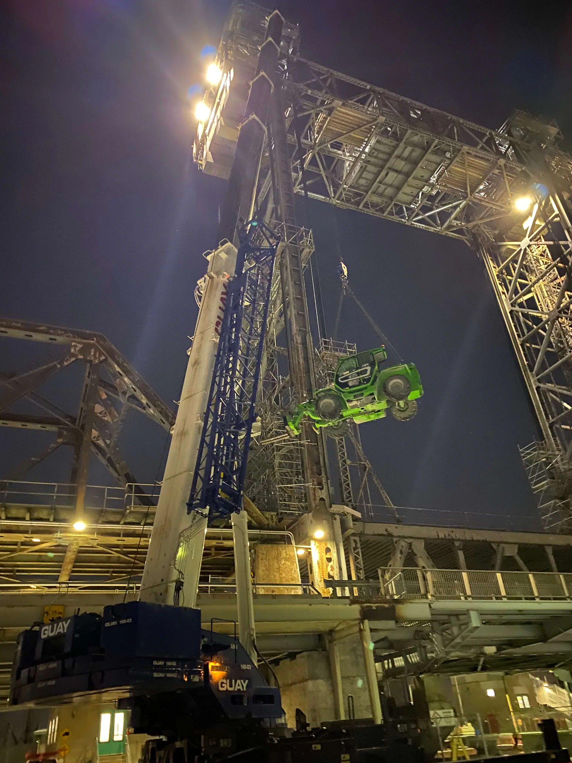 Dramatic night lift of a large green mobile machine suspended inside a tall steel industrial structure, with cranes and floodlights.
