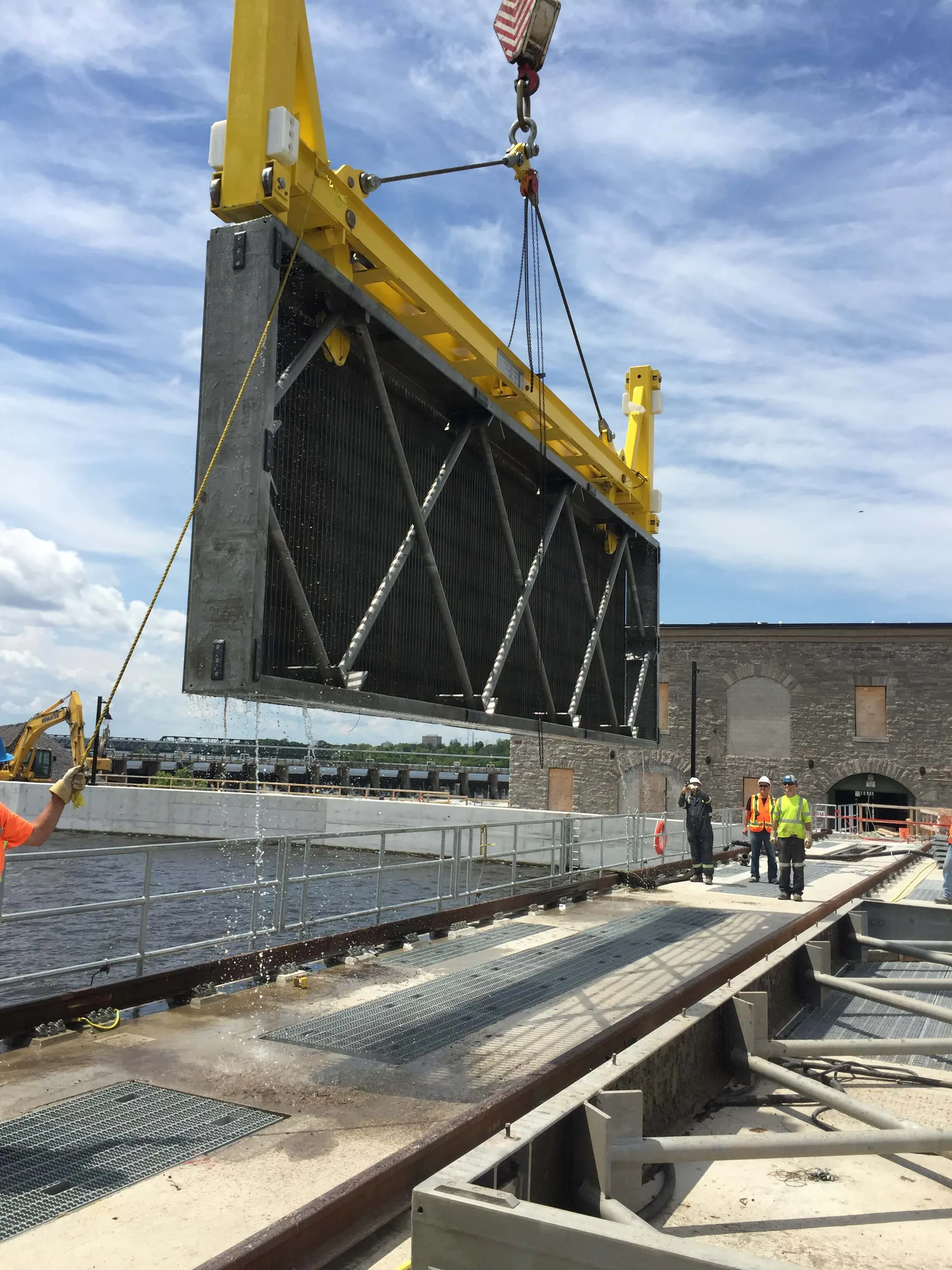 Large black finned rectangular equipment panel hangs from a yellow lifting beam outdoors as three workers guide the operation.
