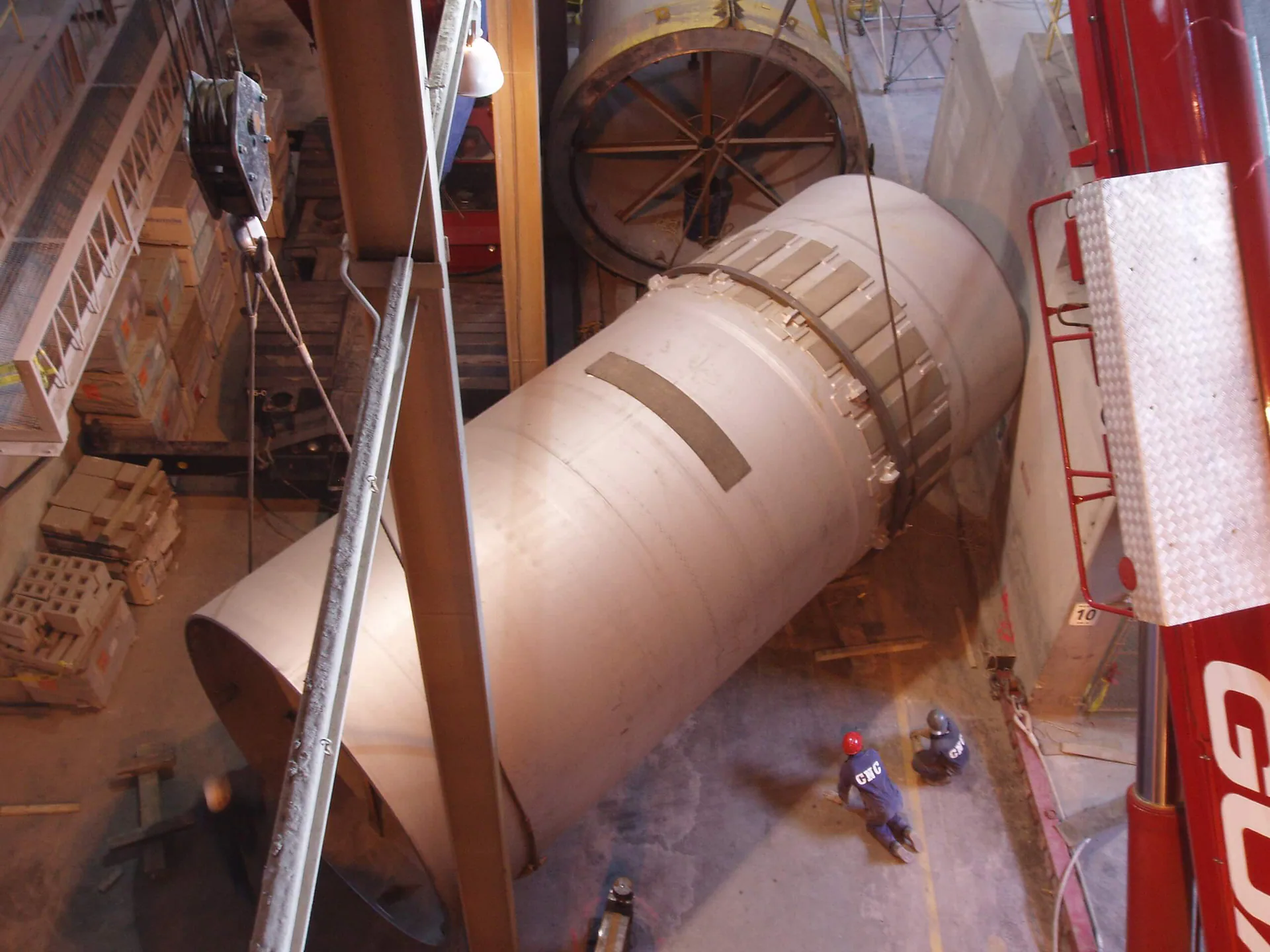 Large cylindrical shell section suspended vertically inside an industrial plant during a crane lift.