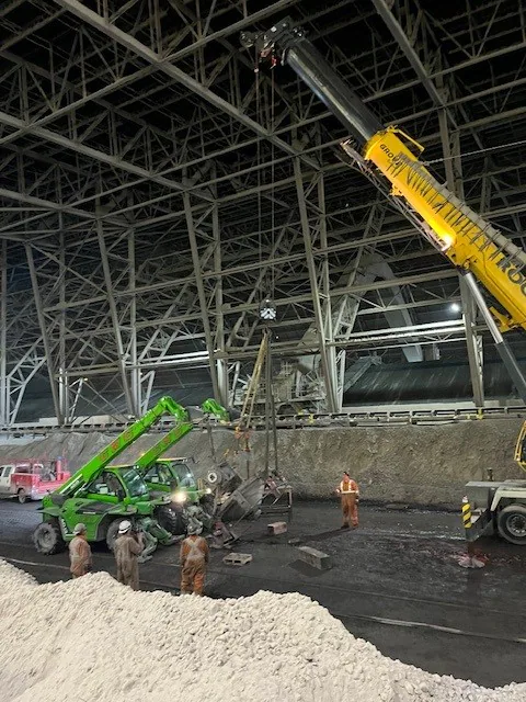Mobile crane and telehandler handling a heavy mechanical component inside a large industrial hall with workers nearby.