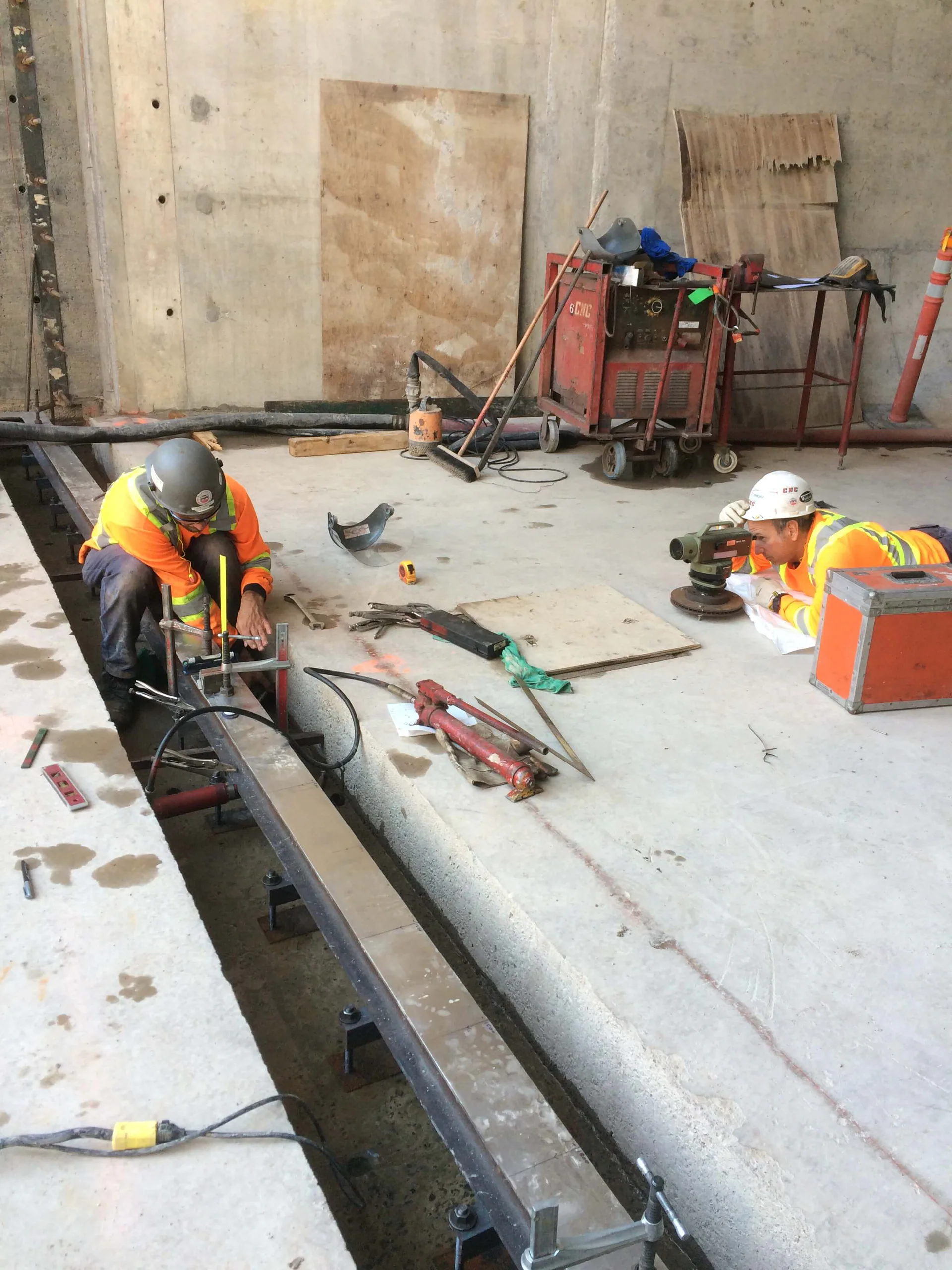 Two CNC-branded workers in PPE precisely measuring and setting a long steel rail or beam beside a floor opening using a surveying instrument.