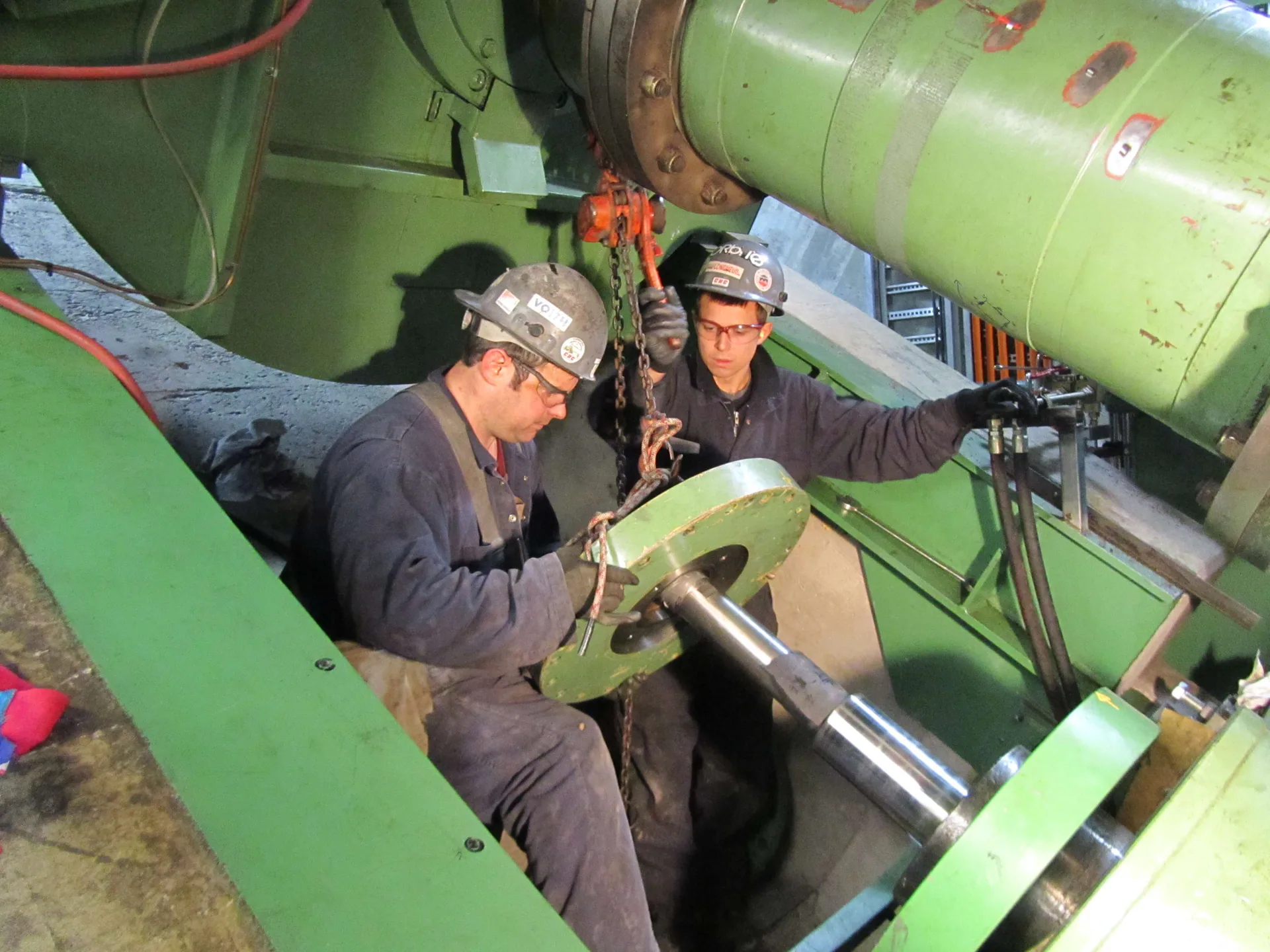 Two hard-hatted workers handle a shaft or bearing component with a chain hoist inside large green rotating machinery.
