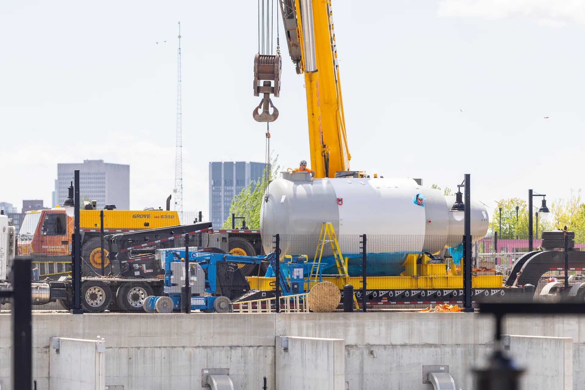 Worker atop a large horizontal cylindrical vessel-like component while a crane hook and rigging hang above it.