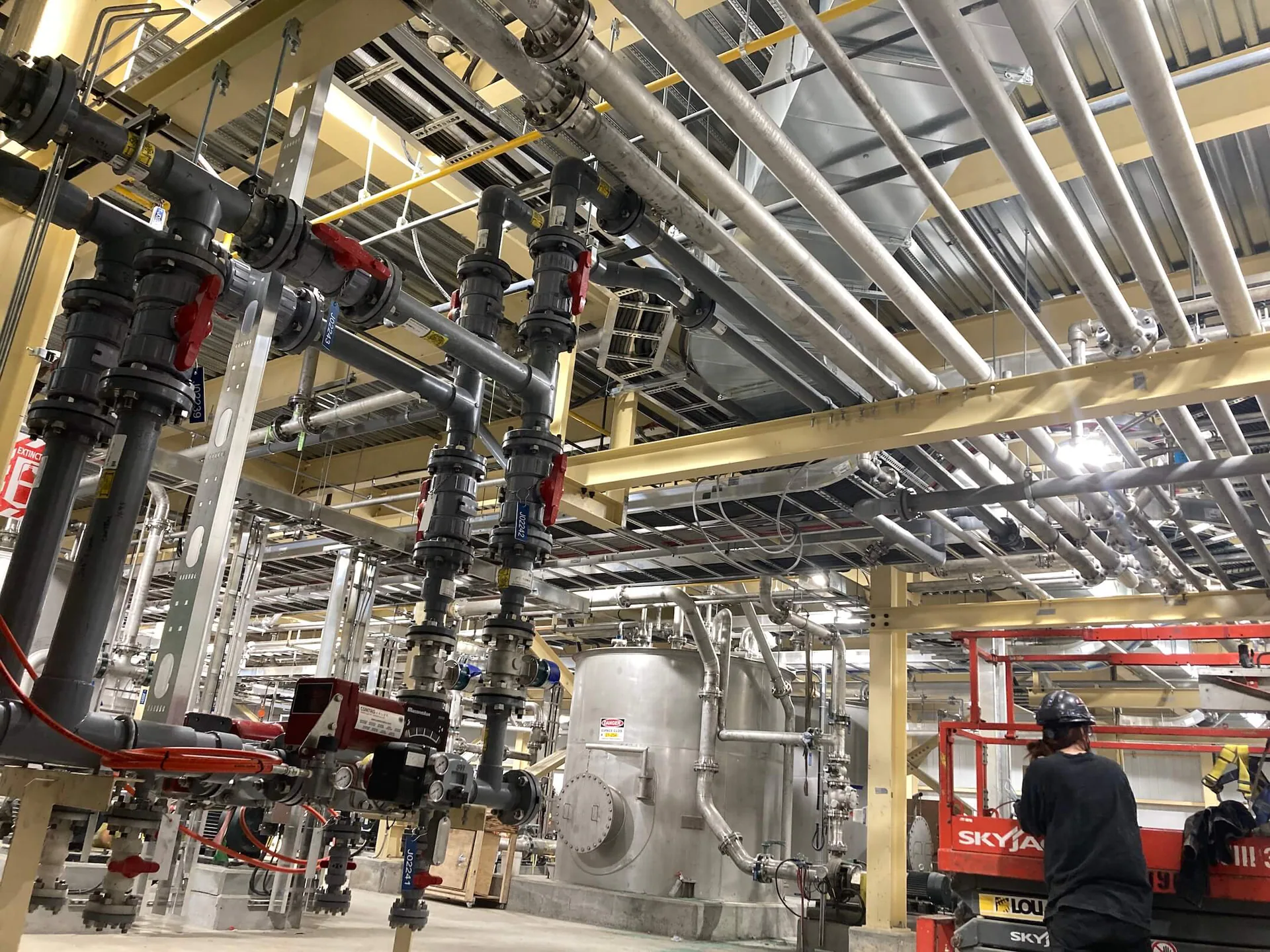 Dense interior process-piping system includes stainless lines, valves, tanks, supports and a worker or lift at lower right.