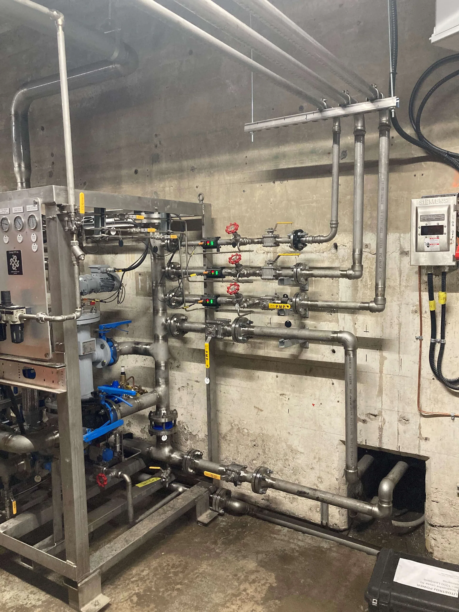 Stainless and small-bore process piping, valves, gauges and a compact equipment skid mounted against a concrete wall.