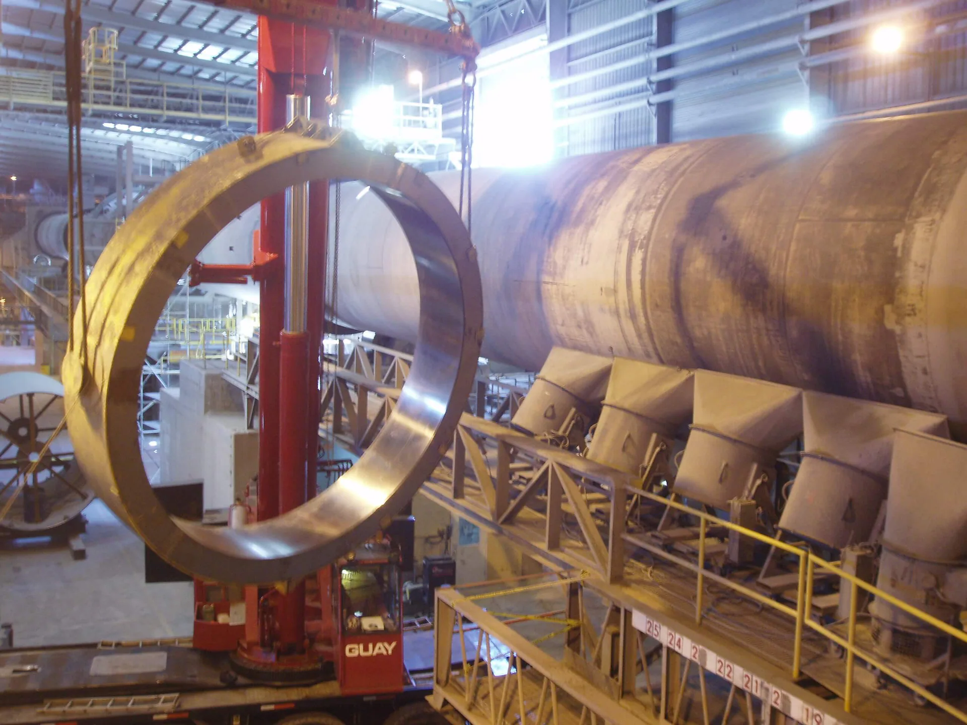 Huge steel ring suspended from a mobile crane beside a long cylindrical industrial shell.
