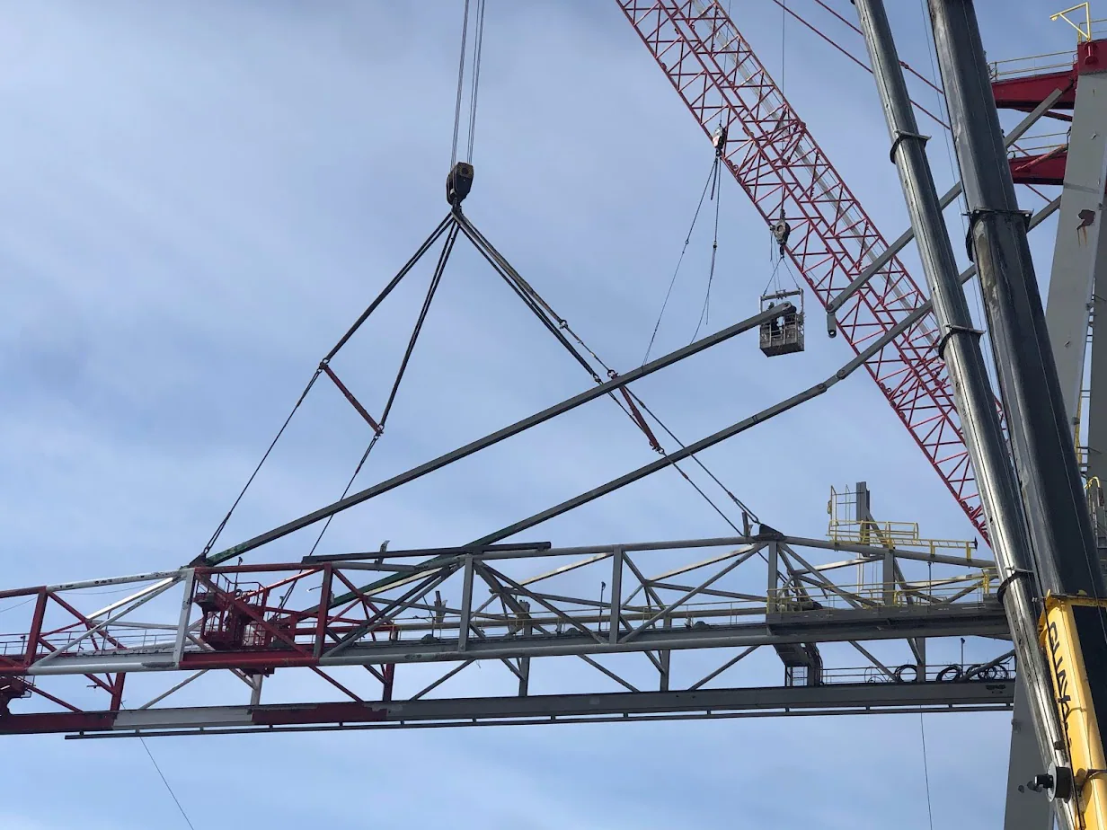 Long industrial truss or boom section hangs from crane rigging beside a stacker-reclaimer-like machine.