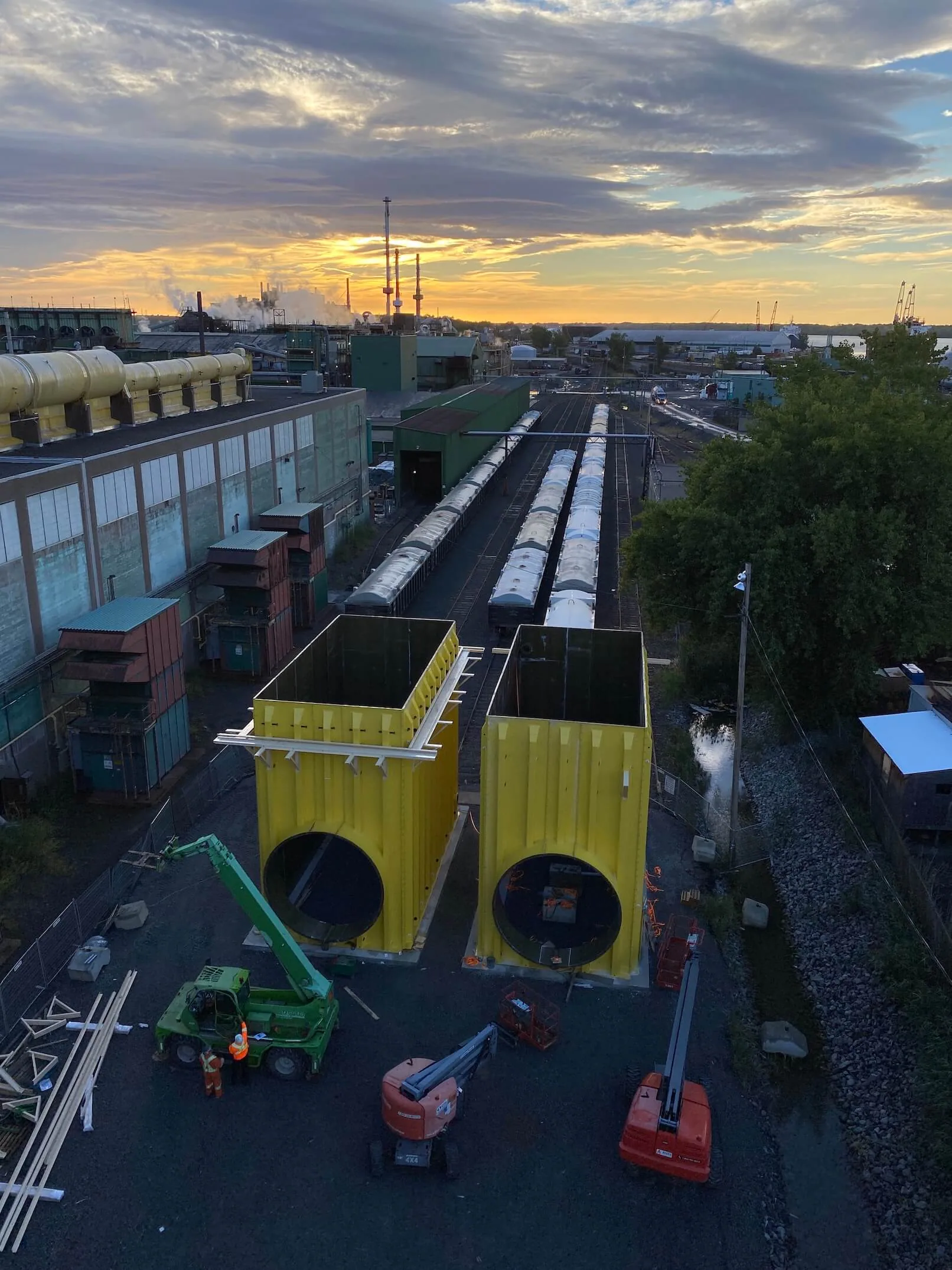 High-angle sunset view of two large yellow open-top steel modules with circular openings, mobile equipment, railcars, and a broad industrial facility.