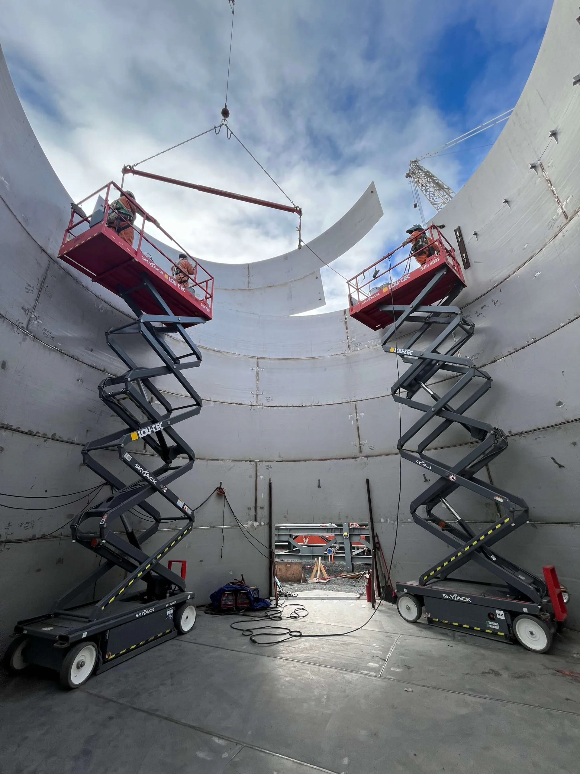 Inside a very large circular metal tank or vessel shell, two workers on scissor lifts guide a curved steel plate suspended from a crane or spreader.
