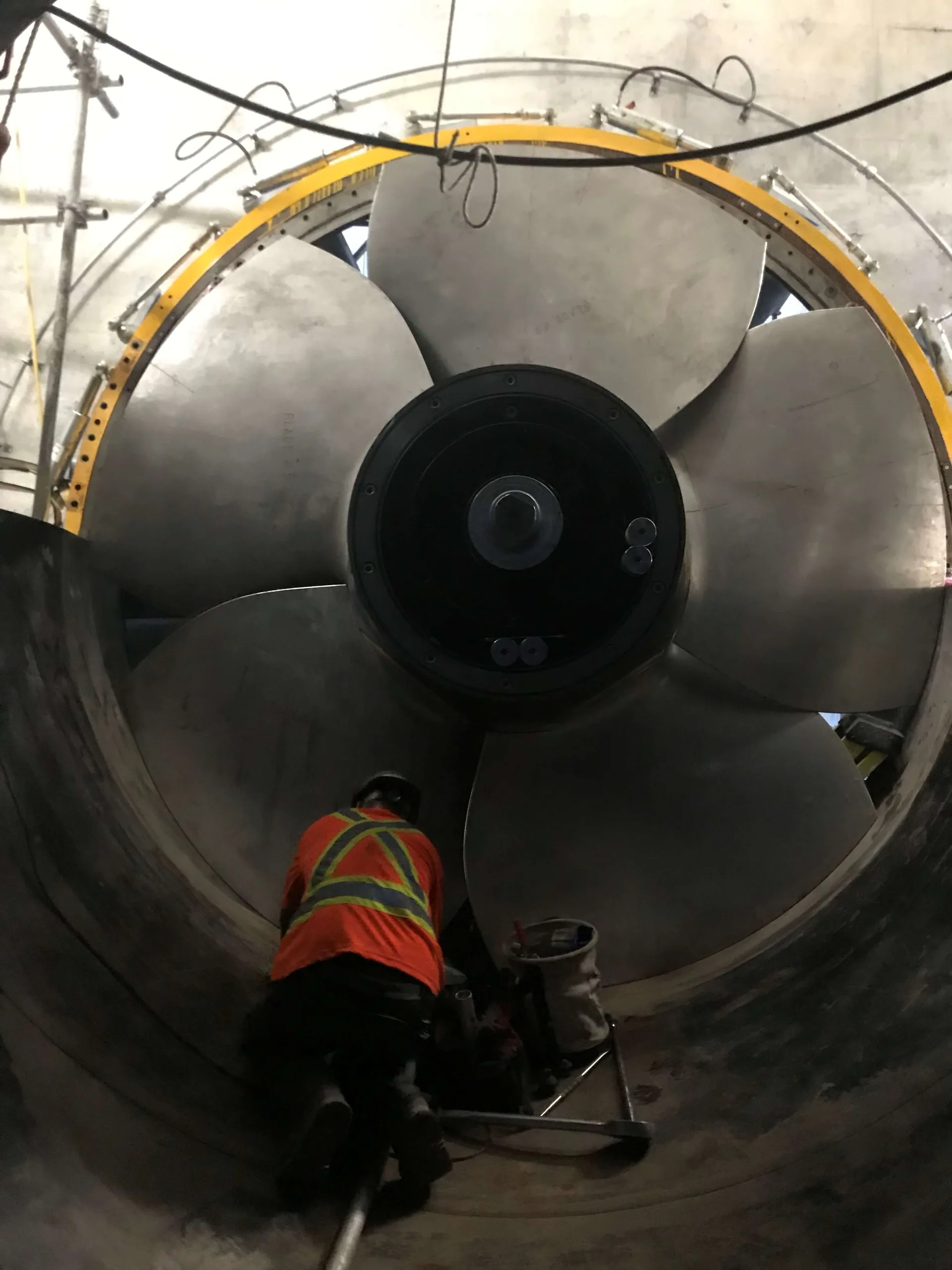 Worker inside a huge circular housing beneath an enormous multi-blade axial fan, propeller, or turbine-runner-like assembly.