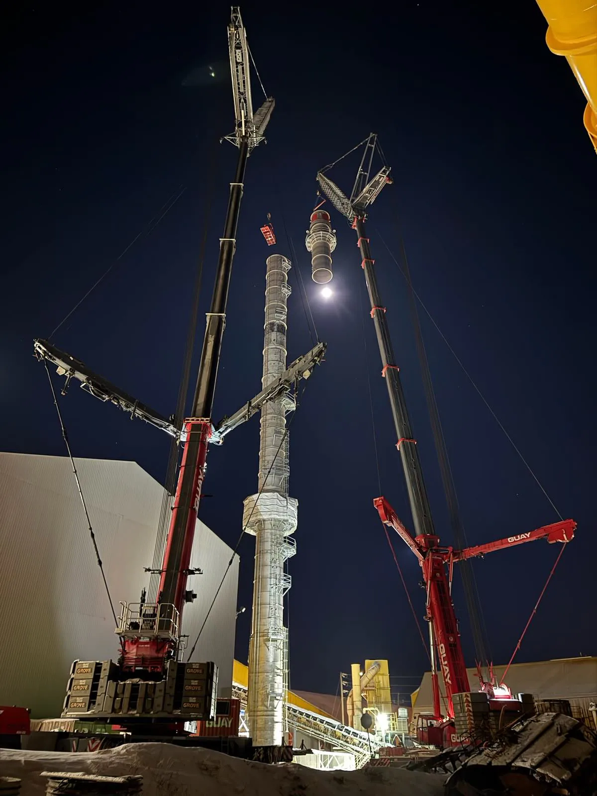 Dramatic nighttime operation uses multiple cranes around a tall industrial stack or column section.