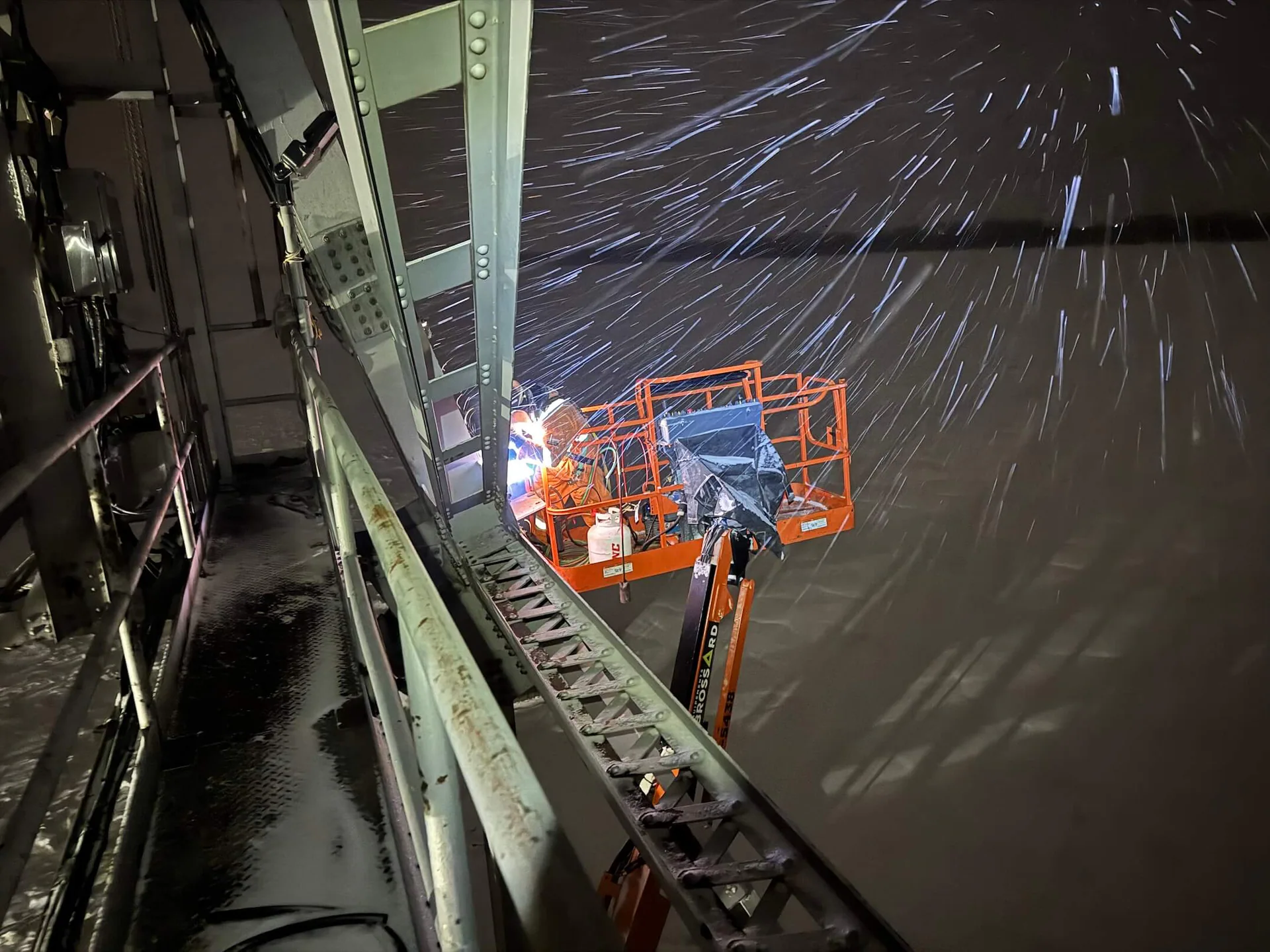 Worker in a boom lift welding or cutting a large steel bridge member at night in winter, with visible sparks.